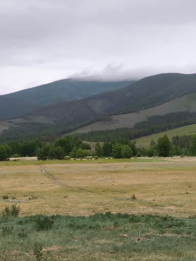 The image depicts a panoramic view of a vast, open landscape with rolling hills and mountains under a cloudy sky. In the foreground, a dry, yellowish-brown field is visible, with patches of green grass and small bushes scattered across it. A faint, winding path is discernible across the field, suggesting human or animal passage. The middle ground is dominated by a dense forest of green trees, with some lighter-colored deciduous trees interspersed. Beyond the forest, the landscape rises into progressively steeper, tree-covered hills, leading to larger mountains in the background. The highest peaks of these mountains are partially obscured by a thick layer of clouds. The overall impression is one of a natural, undeveloped area, possibly in a rural or mountainous region. The location is identified as Kharamodun, Russia.