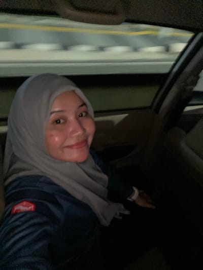 A woman wearing a light grey hijab and a dark blue jacket is captured in a close-up, selfie-style shot from inside a vehicle. She is smiling and looking towards the camera. The background shows a blurred view of city lights and streetlights passing by, indicating the vehicle is in motion, likely on a road or highway in Jawa, Indonesia. The lighting is dim, with some highlights on the woman's face. The interior of the car is visible, including the dashboard and part of the steering wheel.
