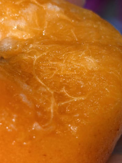 The image is a close-up, macro shot of the textured surface of an orange fruit. The texture appears to be juicy and slightly fibrous, with small air bubbles or water droplets visible on the surface. The lighting highlights the glistening and translucent qualities of the fruit's flesh, with areas of lighter and darker orange indicating the depth and contours of its surface. The background is out of focus and shows a gradient of colors, including purple and dark blue. There is no indication of the city of Orenburg, Russia, or any human presence.