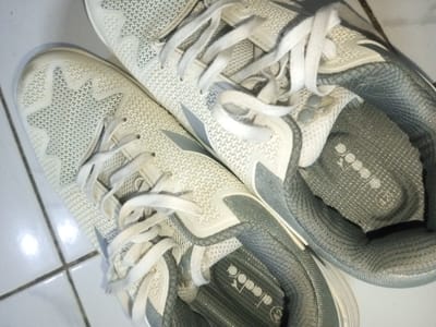 Two white Diadora sneakers sit closely together on a white tiled surface. The sneakers are laced with white laces. A star pattern is visible on the toe of one shoe. The interior of one shoe reveals a gray fabric lining and the Diadora logo with the size "42" imprinted on the insole. Both shoes feature gray accents around the opening.