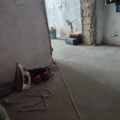 This image depicts an interior scene within a building that appears to be under renovation or construction. The floor is a rough, grey concrete surface. In the foreground, on the left, an electric iron with a white and brown casing is placed on the floor with its braided power cord extending outwards. Next to the iron are two pairs of brown shoes with red accents, one pair clearly showing the word "Polo" in red lettering. A glass jar, partially filled with some substance and bearing a label with red text, sits beside the shoes.
Further into the scene, to the right, a rough-textured brick pillar is visible, suggesting structural elements of the building. Beyond the pillar, the wall is rendered in a rough, light-colored plaster. In the background, to the left of the pillar, a green bucket contains tools, including what appear to be pliers and a hammer. A can of expanding foam with a spray nozzle is positioned next to the bucket. Stacked on the floor behind the can are two toolboxes, one red and black, and another black case. Electrical outlets are visible on the plastered wall in the background. The lighting in the scene appears to be natural light coming from an unseen source, casting shadows on the floor. The image is taken from a slightly elevated angle, looking into the space. There are no people visible in the image. The location specified as Alga, Kyrgyzstan, is contextual information and not directly evident from the visual content of the image itself.
