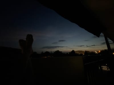 The image depicts a twilight scene from a slightly elevated viewpoint, possibly a balcony or rooftop, in the city of Vénissieux, France. The sky is a deep indigo transitioning to lighter shades of blue and grey, with faint clouds scattered across it. A sliver of light can be seen on the horizon, suggesting the last vestiges of sunset or the first signs of dawn.

In the foreground, silhouetted against the sky, are two feet. The toes are clearly defined, indicating they are bare. The feet are positioned centrally in the frame, and appear to be relaxed, possibly resting.

Below the sky, the cityscape is visible but largely obscured by darkness. Silhouettes of trees and buildings are discernible. Some distant lights are twinkling, hinting at the presence of human activity and infrastructure within Vénissieux. A structure, possibly a railing or fence, runs along the lower right portion of the image, adding a sense of depth and framing the view. The overall mood of the image is quiet and contemplative, capturing a moment of stillness at the edge of day.