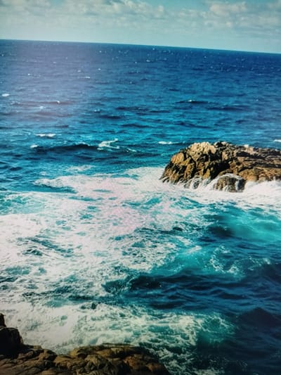 The image shows a view of the ocean with waves crashing against rocks. The sky is partly cloudy, and the water is a deep blue. There are rocks in the foreground and a larger rock formation in the middle ground. The waves are white and foamy. The image does not depict the city of Yashkino, Russia, nor does it contain any people or specific objects.