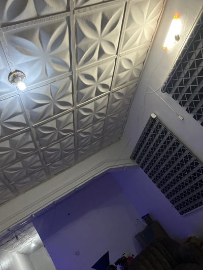 A close-up, low-angle view captures a textured white wall composed of square panels with floral patterns. A light fixture adorns the bottom of the wall. Above, the ceiling features a grid pattern. Part of the adjacent room shows a white wall illuminated by blue light. Furniture and toys are visible in the background.