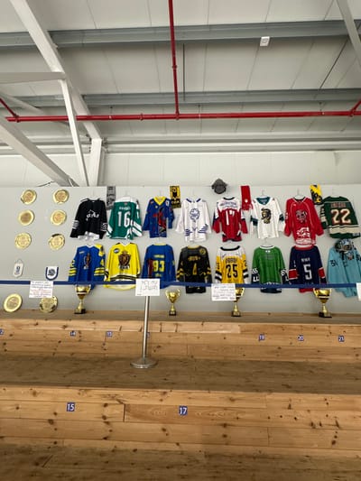 A collection of hockey jerseys is displayed on a wall, each accompanied by a small golden trophy. Below the jerseys are golden medallions and signs. The jerseys showcase various color schemes and team insignias, with visible numbers like 22, 16, 5, 93, and 25. The background consists of a white wall on the left and a wooden plank wall with numerical markings ranging from 15 to 21 on the right. The scene appears to be a hockey memorabilia exhibit.