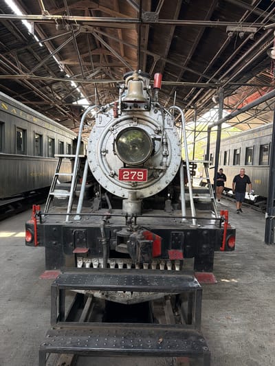 The image depicts the front of a steam locomotive engine, bearing the number "279", indoors at a location in Cuautla, Mexico. The engine is primarily silver and black, with red accents on some components. The locomotive's headlight is prominent in the center. Behind the engine, a train car with gold lettering can be partially seen. Two people are visible in the background, appearing to stand on or near the train car. The building housing the locomotive has a metal framework and what appears to be a corrugated roof. The floor is made of concrete.