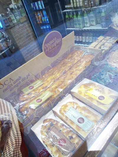 This is a photograph of a bakery display case in Besalú, Spain. The case contains various types of "coca," a traditional Spanish flatbread. In the foreground, there are several long, flat cokes wrapped in plastic, some topped with what appears to be fruit or nuts. The cokes are displayed in cardboard boxes. Behind the cokes, there are shelves filled with bottled beverages, including wine and other liquids. The bakery's signage, in Catalan, is visible on a purple and yellow circular sticker that reads "Coques de Perafita" and "FORN FRANQUESA DES DE 1932." The background shows a brick wall and more shelves of products, suggesting this is a well-stocked establishment. The overall impression is of a busy, traditional bakery.