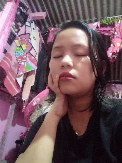 A person with closed eyes rests their face on their hand. They have short, dark hair and wear a black shirt with a delicate gold necklace. The background is filled with various items, including pink accents. A pink fan sits behind the person. The surroundings are cluttered, with items like towels and decorative pieces visible. The overall impression is of someone relaxed in a busy, possibly small, space.