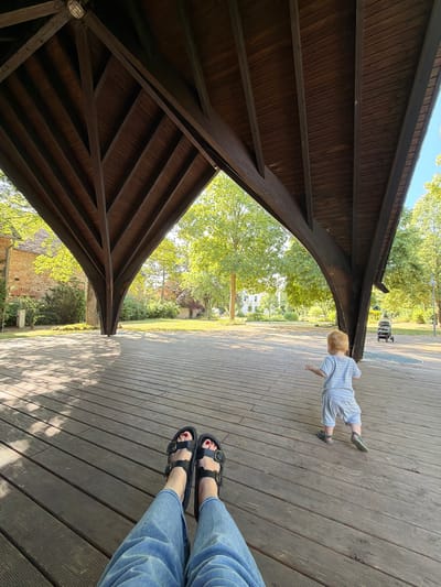The image depicts an outdoor scene beneath a wooden pavilion structure. In the foreground, a person's legs and feet, wearing blue jeans and black sandals, are visible, resting on a wooden deck. The person's toenails are painted red. Further into the scene, a young child with blond hair, wearing a light blue striped outfit, is walking away from the viewer towards a stroller parked in the background. The child is on the wooden deck, which extends into a grassy area with trees. Behind the trees, a building with a stone facade and a tiled roof is visible. The location is identified as the "Bingerbrücker Straße" area in Bad Sobernheim, Germany. Sunlight filters through the trees, casting shadows on the deck.