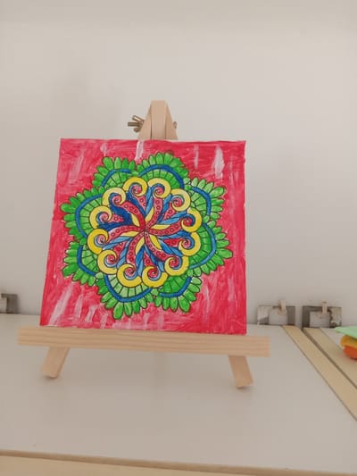 A square, red canvas with a colorful, circular mandala design is displayed on a small, wooden easel. The mandala features intricate patterns in blue, yellow, green, and red, with white highlights. The canvas is positioned on a white surface, possibly a table or desk, against a plain, light-colored wall. In the background, to the right, are two small, grey objects with white cylindrical elements, and a long, light-colored wooden plank. Further to the right, a stack of colorful items, including orange and green, are visible. The lighting suggests an indoor environment with natural light coming from the side. The scene is devoid of any people or specific landmarks that would identify it as being located in Kielce, Poland.
