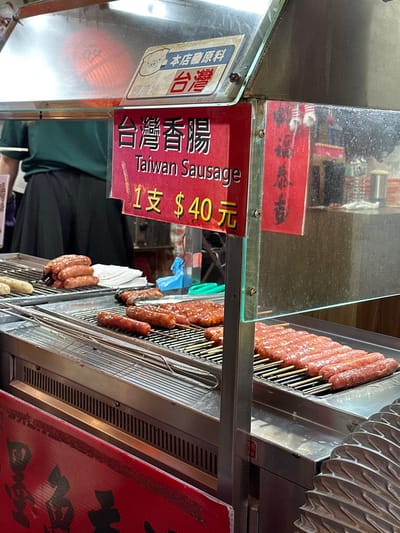The image depicts a street food stall in New Taipei City, Taiwan, specializing in Taiwan Sausage. The exact location within New Taipei City is not determinable from the visual evidence.
The foreground is dominated by a metal-framed food cart with clear acrylic or glass panels forming a protective barrier above the cooking area. A prominent red rectangular sign, centrally positioned, displays "台灣香腸" (Taiwan Sausage) in large white Traditional Chinese characters, followed by "Taiwan Sausage" in smaller English text. Below this, the price is stated as "1 支 $40 元" (1 piece $40 NTD) in yellow characters. Above this sign, a white rectangular sign with a pig icon reads "本店原料 台灣" (This store's ingredients: Taiwan) and "原產地 台灣" (Place of origin: Taiwan). A smaller, vertical red sign with black characters is affixed to the right side of the main sign.
On the metallic cooking surface below the signs, multiple grills hold various stages of Taiwan sausages. On the far left, a stack of cooked sausages rests on a white paper liner. To their right, several individual sausages are on the grill. Further to the right, numerous raw or partially cooked sausages are arranged on skewers across two separate grill sections. Some flattened, reddish-brown meat items, possibly a different preparation, are also visible on the grill. Metal tongs or similar grilling tools are laid across one of the grill grates.
In the blurred background, behind the food stall's transparent panels, a person wearing a dark green shirt and a black skirt or apron is partially visible, facing away from the camera. Various bottles and containers, likely condiments or ingredients, are visible on a shelf behind the person. The overall environment suggests an indoor or covered outdoor setting, consistent with a market or food vendor area.