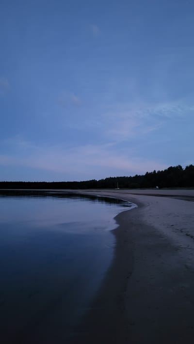 The image depicts a tranquil evening scene at a beach in Zaostrov'e, Russia. The sky is a deep, soft blue, transitioning to a lighter hue near the horizon, with a few wispy clouds scattered across it. A dark line of trees forms the distant shore. The water is calm and reflective, mirroring the twilight sky. The sandy beach curves gently into the frame, meeting the water's edge. A single white structure, possibly a small boat or marker, is visible on the beach in the distance. The overall atmosphere is serene and still, characteristic of dusk.