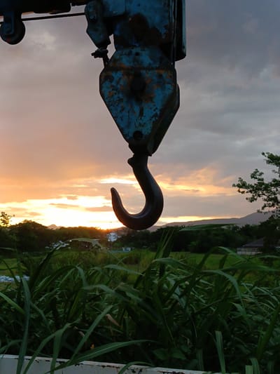 In Ban Patan, Thailand, a close-up shot captures a rusted, blue crane hook hanging against a dramatic sky during sunset. The hook, a central focus, appears weathered with visible signs of age and use. The sky behind it is a gradient of orange and pink hues, indicative of the setting sun, with scattered clouds adding texture. Below the hook, a lush green landscape unfolds, featuring tall grasses in the foreground and a line of trees and distant hills in the background. The overall impression is one of industrial equipment juxtaposed with the natural beauty of the Thai countryside at dusk.