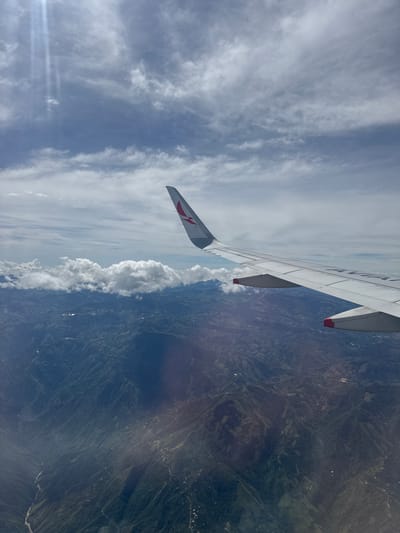 The image, taken from an airplane window, shows a scenic view of mountains and clouds. In the foreground, the wing of the airplane is visible, with a red logo on its tail. The mountains below are covered in lush green vegetation, with some scattered houses and roads visible. A river can be seen winding through the valleys. The sky is partly cloudy, with sunlight streaming through the clouds, creating a dramatic effect. The location is identified as Santa Bárbara, Colombia.