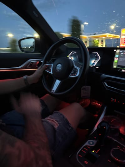 The photo is taken from inside a car, presumably stopped at a Shell gas station. The steering wheel is visible, with a hand interacting with it. The driver is wearing distressed denim shorts. The background shows a Shell gas station visible through the windshield. The image suggests an interior view of a car parked at a gas station at dusk or dawn, with the driver present. The location is not readily discernible as Dragor, Bulgaria.
