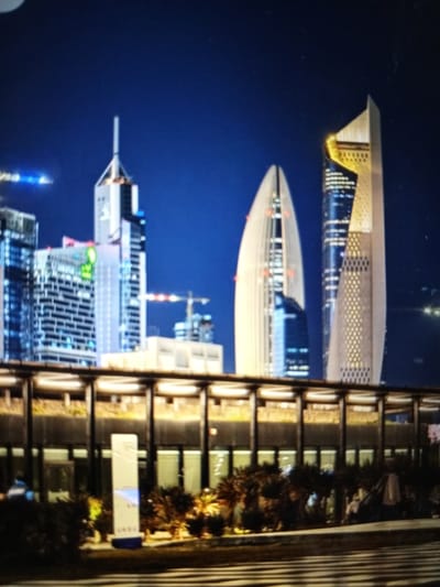 The image depicts a nighttime cityscape with several tall skyscrapers. The buildings are illuminated, showcasing modern architectural designs. In the foreground, there is a low-rise structure with a prominent walkway or canopy supported by dark vertical pillars and horizontal beams. This structure appears to be elevated, with vegetation and some illuminated signage visible below it. The sky is a deep blue, indicative of nighttime. There are no visible people or specific actions taking place in the image, other than the general presence of the illuminated city. The scene is static and focuses on the architectural elements of the urban environment. The location is identified as Golbembal, Nigeria, though this specific geographical context does not alter the visual description of the urban landscape itself.