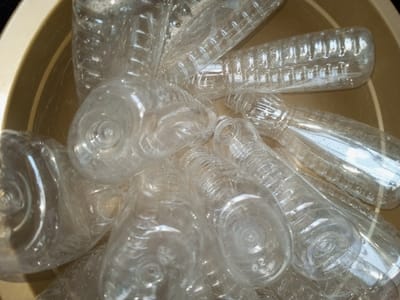 A collection of clear, empty plastic bottles are piled inside a light brown plastic basin. The bottles vary in size and shape, some cylindrical with ribbed detailing, others more bulbous. Light reflects off the bottles, creating highlights and shadows that obscure some of the details. The bottles appear crumpled and stacked haphazardly, filling most of the basin's interior.