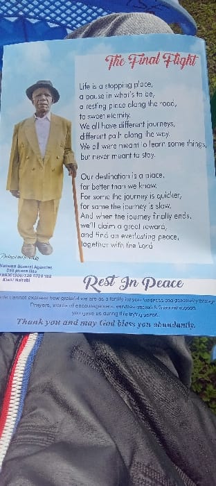 The image displays a vertical memorial program titled "The Final Flight," resting outdoors. The program presents a full-body portrait of an elderly dark-skinned man in a light suit and dark hat, holding a wooden walking stick. Accompanying text includes a reflective poem, "Rest In Peace," and a family's message of gratitude. Contact information for "Wanaru Burial Agencies" notes "Kisii, Nairobi." The program is positioned atop a blue plastic crate, a grey fabric item, and a black garment featuring a red, white, and blue striped zipper. Blurred green foliage is visible in the immediate background, indicating natural ground. The location is in Esise ward, Kisii County, Kenya, as referenced by the program's contact details.