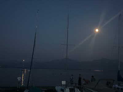 The image depicts a nighttime scene at a marina in Gstadt am Chiemsee, Germany. Several sailboats are moored, their masts silhouetted against the dark sky. One sailboat in the foreground has a blue sail partially visible. A bright moon illuminates the water with a strong glare, casting light rays that streak across the frame. The water's surface reflects some of the moonlight and the lights from the boats, creating shimmering patterns.

In the background, the outline of distant hills or mountains is faintly visible. On the deck of one of the boats, several people are gathered. Their reflections can be seen in the water below, creating an ethereal effect. The individuals appear to be casually dressed and are interacting with each other. The overall atmosphere is peaceful and tranquil, suggesting a calm evening by the lake. The time of day is clearly night, indicated by the darkness and the presence of the moon. There is no visible text in the image.