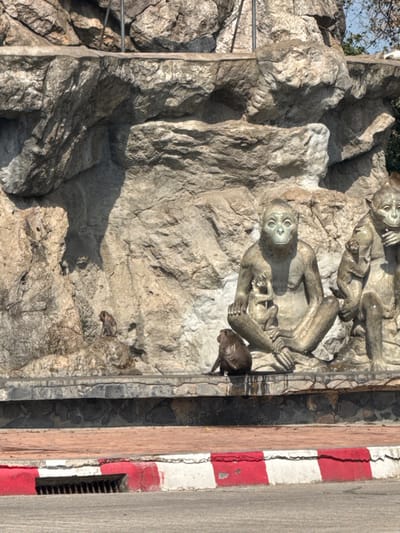 A gray rock face showcases a monkey sculpture with a realistic brown monkey perched nearby. The sculpture features three monkeys in various poses, one holding a baby. A safety railing ascends the left side of the rock. Adjacent to the rock face is a roadside barrier with red and white stripes, indicating a street or road. A section of sidewalk is visible on the right edge.