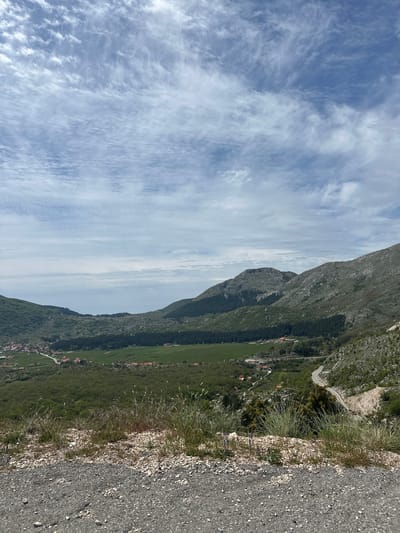 The image captures a landscape scene in Njeguši, Montenegro. A mountainous terrain dominates the view, featuring a series of ridges covered in vegetation. Below the mountain range, a green valley stretches out, dotted with residential buildings and agricultural land. A winding road snakes its way along the mountainside, and a paved road edges the view. The sky is visible above the mountains, showcasing a mix of blue hues and white clouds. The environment appears natural and rural.