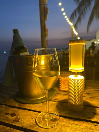 The image depicts an outdoor evening scene at a waterfront establishment in Thành phố Phan Thiết, Vietnam, captured from a low perspective looking across a wooden slatted table. In the immediate foreground, a clear wine glass, approximately half-filled with a pale yellow liquid (likely white wine), stands prominently. To its right, a two-part decorative lamp emits a warm yellow light; its lower section is a cylindrical lantern with a perforated, honeycomb-like pattern, while the upper section is a clear, cylindrical container. To the left and slightly behind the wine glass, a metallic ice bucket holds a dark-colored bottle, partially covered by a light-colored fabric or sleeve, resting on a dark coaster. A small, dark-colored woven basket is visible on the table behind the lamp.
The background reveals a twilight sky, transitioning from deep blue at the top to lighter purplish-orange hues near the horizon line. A body of water is visible to the left, showing faint reflections or distant lights. A string of warm white globe lights extends diagonally across the mid-ground. Silhouetted palm tree fronds are visible on the right. In the far background, blurred figures of people and illuminated structures (likely part of a restaurant or bar) are discernible, along with additional distant lights on the horizon. The overall environment suggests an ambient, relaxed social setting.
