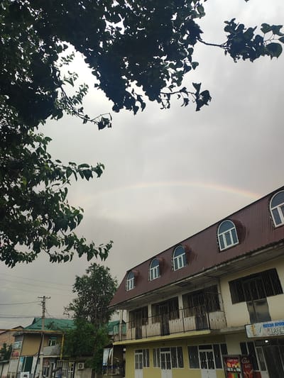 The image depicts an outdoor scene with a partially visible rainbow against a cloudy sky, suggesting recent or ongoing rain. In the foreground, a lush green tree with dark leaves partially obscures the view. A multi-story building with a dark brown, sloping roof and arched dormer windows is visible on the right side of the image. The building has a cream-colored facade with multiple windows and balconies. On the left side, other buildings with green and yellow facades, as well as power lines and a utility pole, are present. The scene appears to be in an urban or semi-urban setting, likely in the city of Tursunzoda, Tajikistan, given the presence of Cyrillic script on a sign that reads "МАГОЗАИ КУРОН" and "АРЗОН." No people are clearly identifiable in the frame. The overall atmosphere is subdued due to the cloudy weather, with the rainbow offering a touch of color.