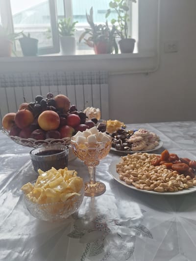 The image shows a table laden with various food items, set in what appears to be a domestic setting, possibly in Novo Stroyka, Kyrgyzstan. The table is covered with a white, patterned tablecloth.
In the foreground, to the left, is a crystal bowl filled with golden, fried pastry, likely a sweet dish. Next to it is a small, dark glass filled with a dark liquid, possibly juice or tea. To the right of the pastry bowl, a delicate, faceted amber-colored glass goblet is filled with white sugar cubes.
Behind these items, a large, ornate glass bowl overflows with a colorful assortment of fruits, including peaches, cherries, and grapes.
Further back on the table, there are several plates with snacks. One plate holds a mix of peanuts and dried apricots and dates. Another plate contains an assortment of chocolates and cookies, some of which are white with red swirls. A small dish with yellow, fluffy confections is also visible behind the fruit bowl.
The background suggests an indoor environment with a window that lets in natural light. Potted plants are visible on a windowsill above a radiator. The overall impression is of a celebratory spread or a gathering with food.