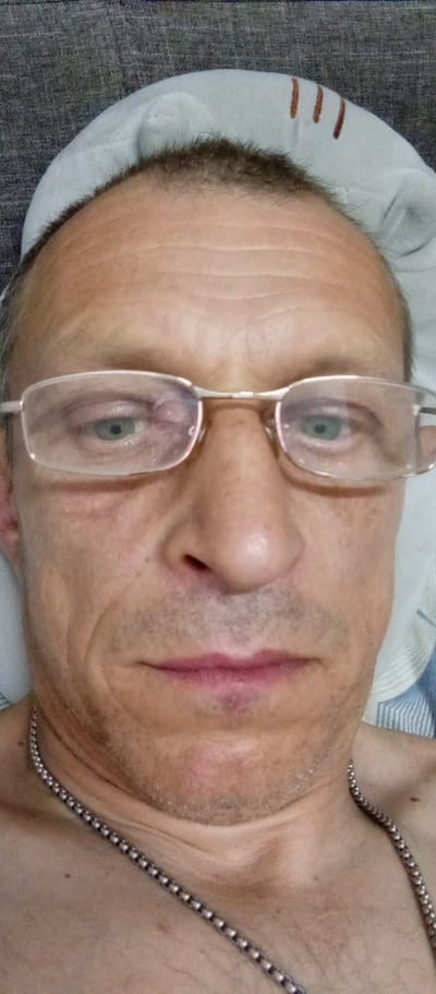 The image is a close-up selfie of a fair-skinned adult male, likely in his 40s or 50s, lying down. He has thinning brown hair, a prominent forehead with visible wrinkles, and light green eyes. He is wearing thin, silver-rimmed glasses. His skin is lightly tanned with some visible pores and a few fine lines. He has a thin, silver chain necklace around his neck, with the beads clearly visible. His lips are slightly parted, appearing natural in color. The background suggests he is indoors, possibly on a bed or sofa, with a gray textured surface visible at the top and a white surface with blue stripes towards the right. There are no other people or discernible objects in the immediate vicinity. The image appears to have been taken from a low angle, with the camera held by the subject. The location within Мастоўскі сельскі Савет, Belarus, is not discernible from the image itself.