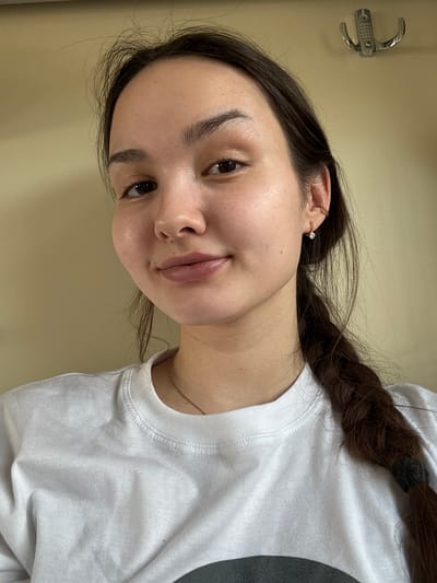 A head-and-shoulders portrait of a young adult female is depicted, positioned indoors in Tayshet, Russia. The subject has dark brown hair, which is parted and pulled back, with a single thick braid resting over her right shoulder, secured with a black elastic. She wears a white crew-neck t-shirt and a thin gold chain necklace. Visible accessories include a small gold hoop earring in the upper cartilage of her left ear and a small gold stud earring with a clear gem in her left earlobe. Her facial features include full lips and dark eyes, with a slight, closed-mouth smile and a direct gaze towards the camera. The background consists of a plain light tan or cream-colored wall, featuring a silver-colored, dual-pronged metal coat hook mounted high on the right side of the frame.