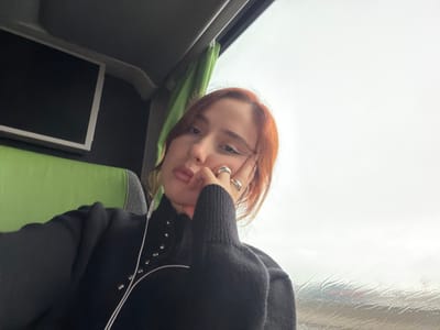 The scene depicts a young adult female seated inside what appears to be a bus or coach, located in Moineville, France. The female subject, with auburn hair, light complexion, and defined eyeliner, faces the camera directly, resting her right hand, adorned with two silver rings, against her jawline. Her lips are slightly pursed. She is wearing a dark, long-sleeved top featuring a high neck, silver stud embellishments along the chest, and a contrasting light-colored curved trim. White wired earphones are visible, with one earpiece in her left ear. To her left, a lime-green upholstered seat is visible, with a dark, unlit screen featuring a white bezel mounted on the wall behind it. To her right, a large window shows an overcast sky and a wet exterior, indicative of rain, with blurry red and white shapes, possibly distant vehicle lights, faintly discernible through the water-streaked glass. A folded green curtain is visible at the upper right edge of the window frame. The interior ceiling is dark gray or black.