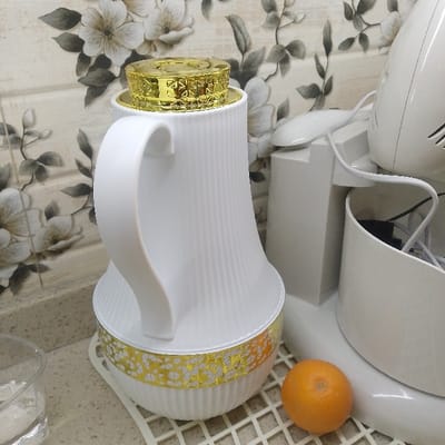 A white carafe with a gold top and base sits on a white, grid-patterned surface. Next to it is a white stand mixer, a small orange is placed near the mixer base. A partially visible glass of water sits in the foreground. The backdrop features floral wallpaper in shades of gray and white.