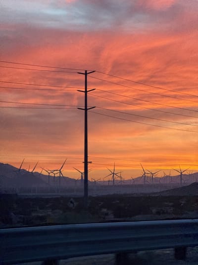 The image captures a landscape with several wind turbines in the city of Desert Hot Springs, California, United States. The turbines are in the background, silhouetted against a vibrant sunset sky with shades of orange, pink, and grey clouds. An electrical power pole and overhead lines are prominent in the foreground, partially obscuring the view of the turbines. A dark horizontal line, likely a road or barrier, runs along the bottom of the frame.