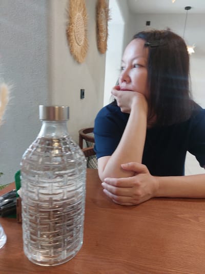 The image depicts a woman sitting at a wooden table, looking away from the camera with her chin resting on her hand. She is wearing a dark blue shirt. To her left, on the table, is a clear glass bottle with a textured pattern, filled with a clear liquid, likely water. In the background, on the wall, are two decorative woven circular objects and a light fixture. The overall setting appears to be an indoor cafe or restaurant. No specific geographical markers for Kyzylorda, Kazakhstan are visible in this image.