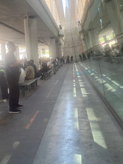 This media file depicts a group of people in an outdoor, covered walkway, possibly a bus station or a transit hub, in Parakar, Armenia. The scene is characterized by a long, reflective glass wall on the left, which mirrors the sky and surrounding buildings, suggesting it is daytime. Sunlight filters through an overhead structure, casting distinct, elongated shadows on the concrete floor, indicating a bright and sunny day.
The subjects are a diverse group of individuals, appearing to be in a queue or waiting area. Most are standing, some are seated on benches, and a few are in motion. Their attire ranges from casual to slightly formal. The people are positioned along a long, tiled walkway with metal benches lining the right side. Some individuals are looking at their phones, while others appear to be conversing or simply waiting. The overall mood seems to be one of patient waiting, with no obvious signs of distress or excitement.
Notable details include the architectural style of the walkway, with its modern, clean lines and extensive use of glass and concrete. The shadow patterns are a prominent visual element, adding a dynamic quality to the scene. There is no visible text in the media file. The location is identified as Parakar, Armenia, which provides a geographical context for the image. The reflection in the glass wall hints at an urban or semi-urban environment with some greenery visible, suggesting the presence of trees or other vegetation.