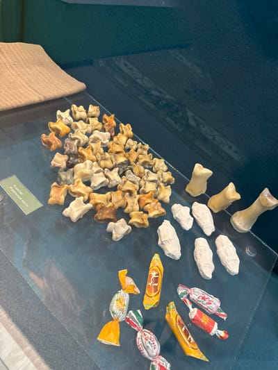 The image displays a collection of ancient artifacts and modern candy, likely within a museum exhibit in Fort-Shevchenko, Kazakhstan. In the upper left corner, a piece of coarse, tan fabric is draped over an unseen object. The majority of the frame is occupied by a glass surface, upon which various items are arranged.

In the center left, a large pile of small, light-colored bones, appearing to be animal astragali (ankle bones), are scattered across the glass. These bones are varied in shape and color, ranging from off-white to tan and brown, suggesting they are ancient. A small green label with text in a Cyrillic script, likely Russian or Kazakh, is placed among the bones.

To the right of the bones, three light beige, foot-shaped objects, possibly replicas or casts of ancient footwear or bone fragments, are positioned in a row. Below these, a cluster of rough, white, oblong objects, resembling dried or mummified objects, are arranged.

In the foreground, several pieces of colorful, foil-wrapped candy are scattered on the glass surface. One candy wrapper features an image of a horse and rider, another depicts oranges and greenery, and others have red and white striped patterns. The background consists of a dark, teal-colored surface, possibly the interior of a display case. The overall lighting is even, highlighting the details of the objects.