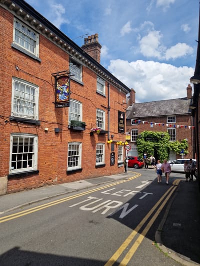 The image depicts a street scene in Knutsford, United Kingdom, likely on a sunny day with scattered clouds. The street is paved, with double yellow lines indicating parking restrictions and painted white markings on the road that read "LEFT TURN".
On the left side of the street is a two-story brick building with white-framed windows. A pub sign, "THE ANGEL", with stained glass depicting an angel, hangs from the building. There are also window boxes with plants and flowers.
On the right side of the street, another building is visible, covered in ivy, with windows and a sign that reads "Antiques Centre". A white car and a red car are parked on the street. Several people are walking on the pavement and crossing the street. The architecture suggests a historic town center.