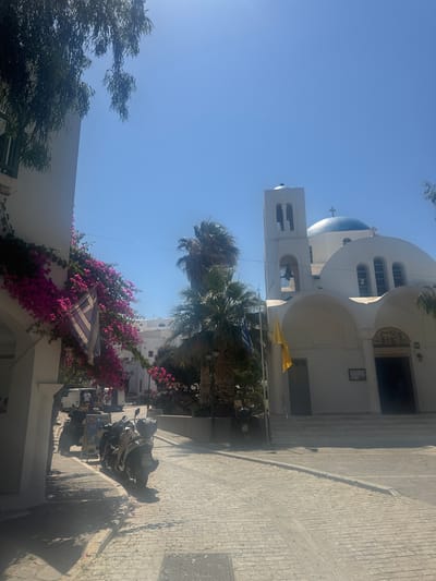 In Naousa, Greece, a sunlit scene unfolds on a cobblestone street in front of a whitewashed church. The church features a striking blue dome topped with a cross, and a tall bell tower. Archways lead to the church's entrance, and steps ascend to the main doors. A Greek flag and a yellow flag hang near the entrance. Bougainvillea in vibrant pink cascades down a nearby white building, alongside a Greek flag with blue stripes. Palm trees, including a tall, slender one, are scattered throughout the scene. Several motorcycles and a quad bike are parked on the street. The sky is a clear, bright blue, indicating a sunny day.