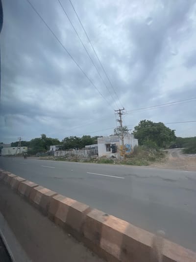 The image depicts a roadside scene in Bethamcherla, India. A paved road runs horizontally across the frame, with a concrete barrier on the left side. To the right of the road, there are several low-rise buildings, some appearing incomplete, along with scattered debris. A prominent electrical pole with wires is visible, and beyond that, a dense cluster of green trees. The sky is overcast with heavy, gray clouds. A person on a bicycle is seen in the distance on the left side of the road.