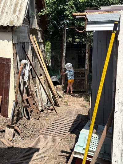 In the city of Yugo-Severnaya, Russia, an outdoor scene unfolds in what appears to be a cluttered backyard or workshop area. A young child, wearing a wide-brimmed hat and orange shorts, stands with their back to the camera, looking into an open doorway of a weathered wooden structure. The doorway reveals a dimly lit interior with what looks like caged sections.

The immediate foreground is dominated by a collection of weathered metal and wooden objects, including a shovel, a rusty gear mechanism, and various planks of wood leaning against a corrugated metal structure. Two metal grates are laid out on the dirt ground, casting shadows. To the right, a tall, yellow pole with black accents leans against a gray wooden structure. Beyond the child and the doorway, more greenery and fencing are visible, suggesting a somewhat overgrown environment. The overall impression is one of disarray and utilitarian outdoor space.