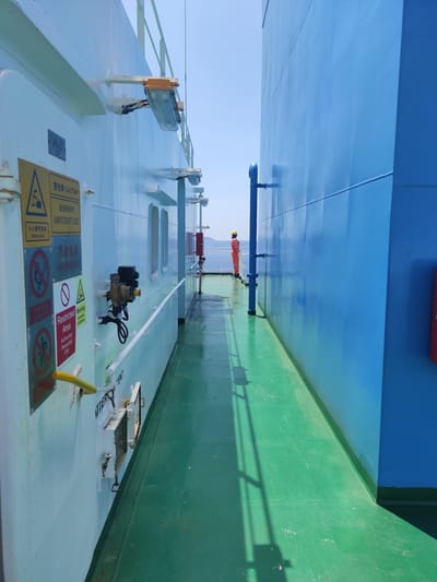 This vertically oriented image captures a working deck of a large vessel, likely a ship or barge, under bright daylight in Kunak, Malaysia. The scene is dominated by distinct colors: a vibrant blue wall or hull section on the left and a contrasting green deck or side wall on the right. The main deck area, visible in the foreground, is painted a light grey or white.

In the mid-ground, a lone individual is visible, dressed in orange coveralls and a yellow hard hat, standing near a blue pipe that runs horizontally across the vessel. The person appears to be performing a task or inspecting the equipment. The vessel's deck features various industrial components, including pipes, valves, a control box with wires, and several hatches or vents. Numerous safety signs are affixed to the deck, providing warnings and instructions.

Notable details include the clear blue sky and strong shadows cast by the ship's structures and the railings on the green surface, indicating sunny weather. Visible text on the safety signs includes "小心酸性电池" (Caution Battery acid), "Battary acid," "警告牌 CAUTION" (Warning sign CAUTION), "总保持关闭" (Always keep closed), "ALWAYS TO BE KEPT CLOSED," "Restricted Warning Area Authorized Personnel Only," and "Comosive" (likely referring to "Corrosive"). Another partial word, "VENT," is also discernible. The overall impression is one of an active industrial environment at sea or in port.