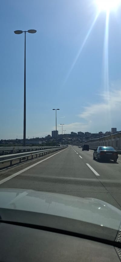 A vertical shot captures a bright, sunny day with a clear blue sky dominating the upper portion. The sun, intensely bright, is positioned in the upper right corner, casting a strong glare and creating lens flares. The lower part of the image shows a view from inside a vehicle, looking out over a highway that curves to the right and recedes into the distance. The highway has multiple lanes separated by white lines. Guardrails line the left side of the highway, and a concrete barrier with decorative yellow and white elements is visible on the right.

In the background, a cityscape unfolds on a hill, featuring various buildings, including tall apartment blocks and smaller houses. Several tall streetlights, each with two horizontally oriented lamps, are spaced along the highway and in the distance.

On the highway, a dark-colored sedan is in the right lane, and a dark van is in the lane to its left, both moving away from the camera. The hood of the vehicle from which the photo is taken is visible in the foreground, reflecting the sky and highway.

The location is identified as the highway in Surakhany Raion, Azerbaijan, based on the context provided.