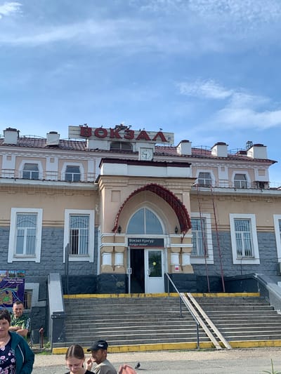 The image depicts the exterior of the Kungur railway station in Kungur, Russia, on a bright, sunny day with a partly cloudy sky. The station building is a multi-story structure with a light beige facade and a dark red tiled roof. Large red Cyrillic letters spelling "ВОКЗАЛ" (VOKZAL, meaning railway station) are prominently displayed on the upper part of the building, along with a clock. Below the main sign, smaller text reads "Вокзал Кунгур" and "Kungur station".
The entrance to the station is accessed via a wide set of stairs with yellow markings on the edges, and a ramp for accessibility. The lower part of the building is faced with dark grey stone. Several people are visible in the foreground, including a man in a green t-shirt looking towards the camera, a woman in a floral top, and a young woman wearing glasses. To the left of the entrance, a map titled "ТУРИСТСКАЯ СХЕМА города Кунгура" (Tourist Map of the city of Kungur) is displayed on the wall. A ladder leans against the side of the building near the entrance. Pigeons are present on the roof and on the ground.