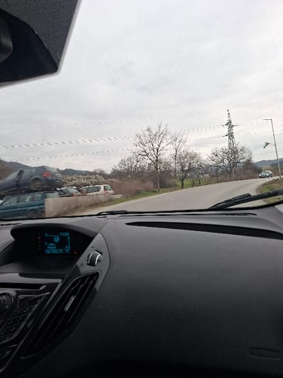 The image depicts a forward-facing view from the interior of a vehicle on a paved road in Cherniche, Bulgaria, under an overcast sky. The vehicle's dashboard, infotainment screen displaying digital information (including "12:51"), and windshield wipers are visible in the foreground. To the left of the road, a vehicle storage or junkyard contains multiple cars, including a dark blue sedan stacked atop a teal minivan, with other vehicles partially obscured behind a low concrete barrier. The road ahead curves gently to the right and appears to ascend slightly. To the right of the road, leafless deciduous trees and sparse dry vegetation line the shoulder. A metal utility pole supporting power lines is visible, along with a street light pole from which a Bulgarian flag is displayed. A dark-colored sedan is visible driving in the distance on the curved road. Distant hills or mountains are discernible on the horizon to the left. No individuals are visible.
