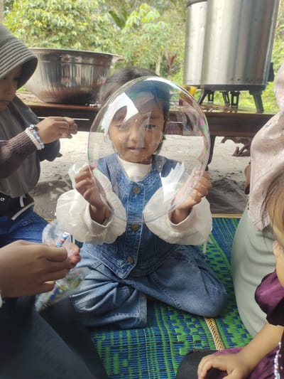 This image depicts an outdoor scene, possibly in Cintadamai, Indonesia, where a group of young girls are engaged in blowing bubbles. In the foreground, a girl wearing a denim jumpsuit is holding a large bubble, looking directly at the camera. Her hands are positioned to keep the bubble intact. To her left, another girl wearing a headscarf and a brown sweater is focused on handling some bubble-making supplies. To the right of the main subject, a portion of another girl wearing a light pink headscarf is visible, as well as a glimpse of a fourth girl in a purple dress. The environment suggests a casual gathering, with natural foliage visible in the background, and large metallic containers and wooden furniture also present. The girls are sitting on a woven mat.