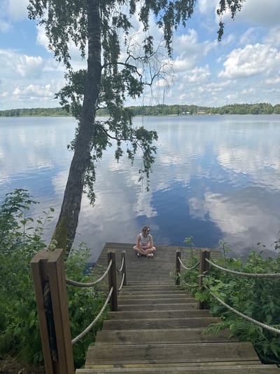 In the city of Priedesmuiža, Latvia, a young woman sits cross-legged on a wooden dock at the edge of a calm lake. The dock features a set of wooden stairs leading down from it. A large tree with leaves overhangs the dock and lake from the left side of the frame. The lake's surface reflects the sky and clouds, creating a serene atmosphere. Greenery and foliage surround the dock and stairs, adding to the natural setting.