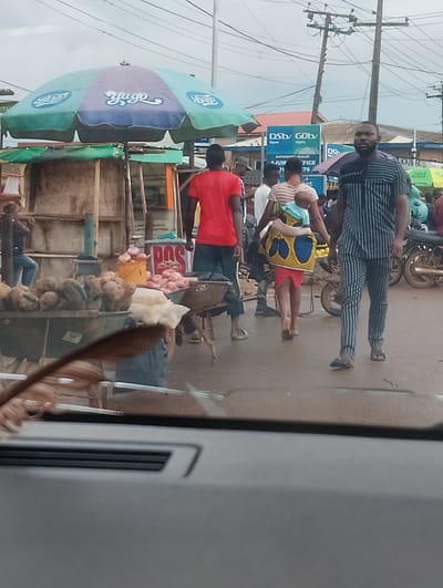 The image shows a street scene in Aketu-Oja, Nigeria. A man in a striped shirt and pants walks towards the camera. Behind him, a woman carries a baby on her back, and further in the background, people are gathered around market stalls. One stall has a large umbrella with "Yugo" written on it. There are also signs for DStv and GOtv. The ground appears wet, suggesting it may have rained recently. The overall impression is of a busy market day in a Nigerian town.
