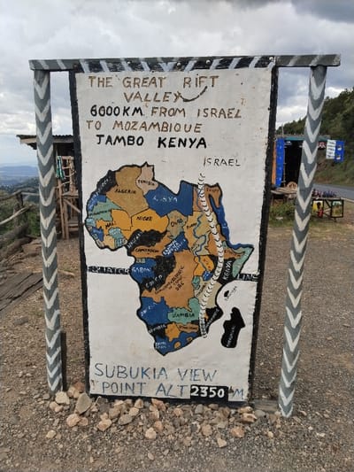 The image displays a hand-painted informational sign at a scenic overlook, identified on the sign as "SUBULIA VIEW POINT ALT 2350M," situated in Waseges ward, Kenya. The central object is a large, rectangular white sign board, vertically oriented, supported by two wooden posts adorned with alternating grey and white chevron patterns.
The upper portion of the sign features the hand-drawn black text: "THE GREAT RIFT VALLEY 6000KM FROM ISRAEL TO MOZAMBIQUE JAMBO KENYA." Below this, a multi-colored map of Africa is depicted, showing various countries, with a prominent wavy white line, representative of the Great Rift Valley, traversing the continent from north to south, extending conceptually from "ISRAEL" (labeled to the right) to Mozambique. The lower section of the sign contains blue text stating "SUBULIA VIEW POINT ALT 2350M."
The sign is positioned on a loose gravel and dirt surface. To the left, a wooden railing and a rustic wooden structure with a corrugated iron roof are visible, overlooking a vast, distant valley. To the right, a paved road runs parallel to the scene, bordered by another makeshift stall-like structure, blue in color with a corrugated iron roof, displaying various items, including small bottles and a framed picture, on an adjacent wooden surface. A lush green hillside forms the background beyond the road. The sky above is overcast with grey clouds. No individuals are present in the scene.