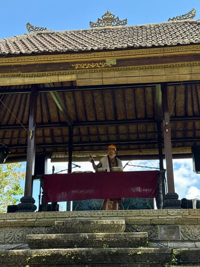 The image, taken in Carangsari, Indonesia, shows an outdoor stage constructed of dark wood and stone. The roof is made of woven material and decorated with intricate gold and silver designs. A red curtain hangs at the back of the stage, obscuring the background. A man wearing a yellow hat and glasses is seen on stage, holding an object. A microphone stands in front of him. Stone pillars with carvings are visible on the right side of the stage. The sky is visible through the gaps in the roof.