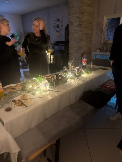 A slightly tilted view depicts a partially consumed feast. A long table covered with a white cloth is adorned with string lights, empty dishes, and liquor bottles. Two women are visible; one holding a drink and the other standing further back. A stone wall with a fireplace, television, and a large clock adorns the background. The scene suggests a lively gathering or party.